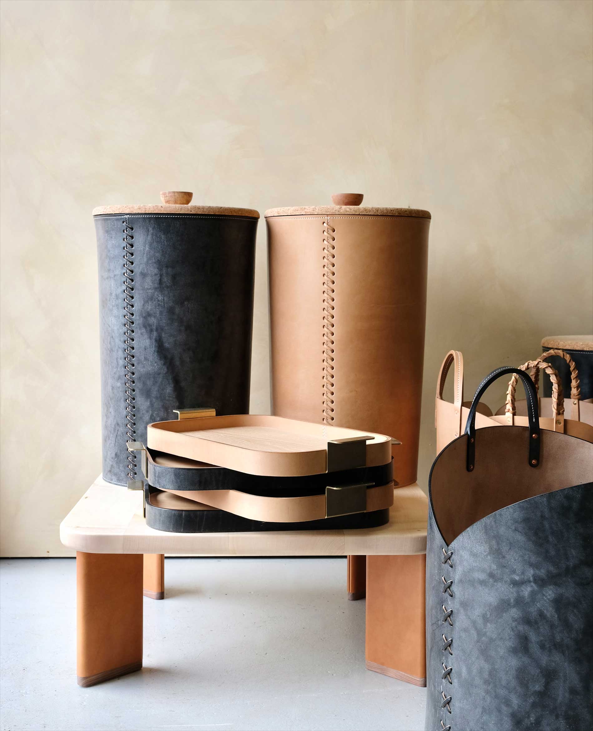 A collection of modern leather home goods, including tall cylindrical containers, nesting trays with handles, and a storage basket, displayed on a light wood table against a neutral background.