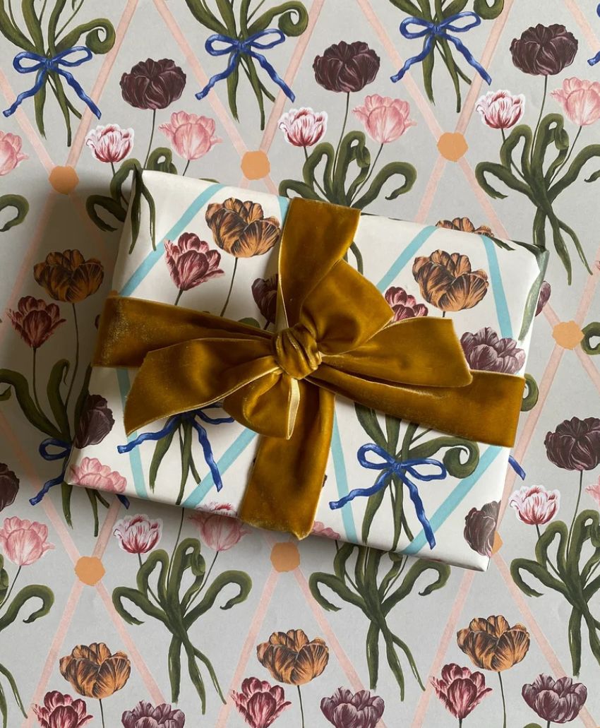 A gift wrapped in floral-patterned paper with a large golden-brown velvet ribbon, placed on matching floral-patterned wrapping paper.
