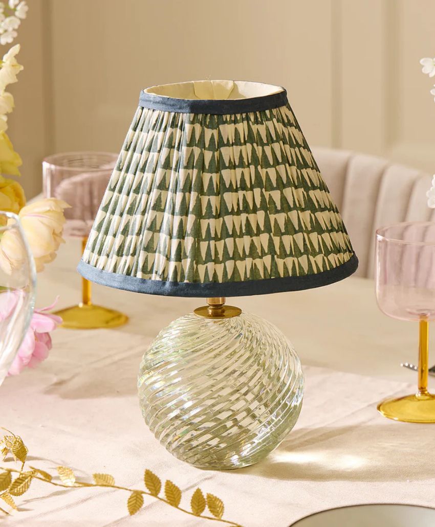 A small table lamp with a round, textured glass base and a patterned fabric lampshade featuring green and white triangular shapes, placed on a light-colored tablecloth.