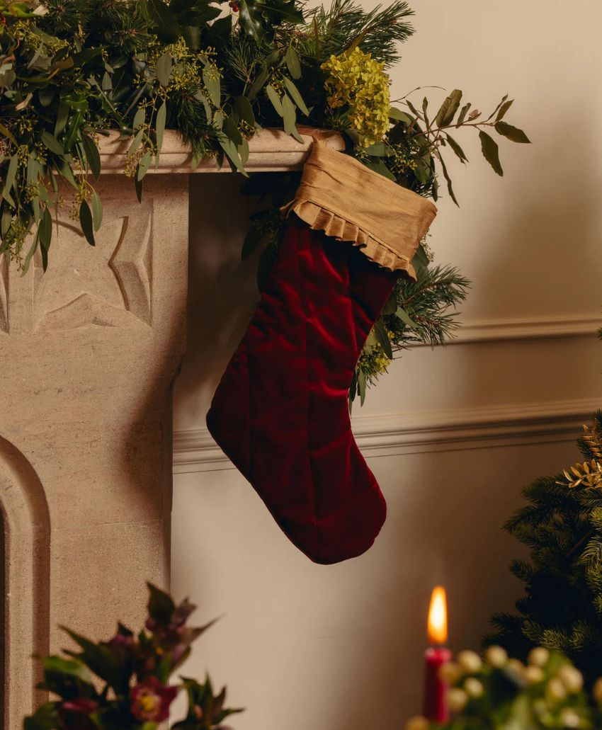 A red Christmas stocking with a tan cuff hangs from a mantel decorated with green foliage; a candle glows in the foreground.
