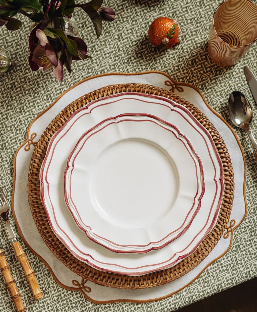 A place setting with three plates of varying sizes, stacked on a woven placemat atop a patterned tablecloth, with a fork, knife, spoon, pink glass, and a decorative orange visible.