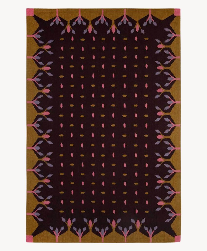 A rectangular rug with a dark maroon center, surrounded by a golden brown border. The edges feature symmetrical, abstract floral patterns in pink, lavender, and black. Pink squares accentuate the corners.