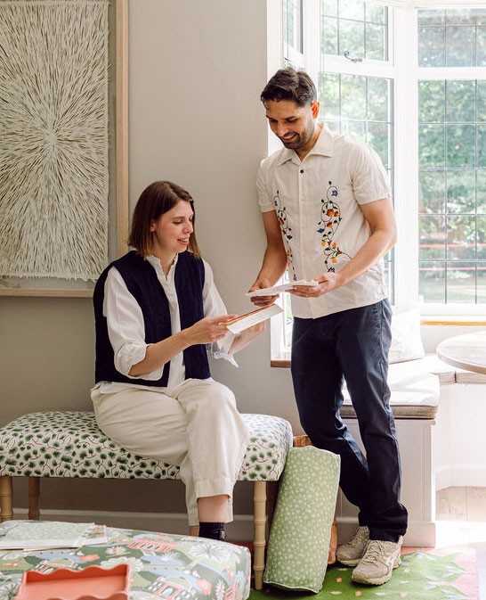 A woman sitting on a bench and a man standing beside her are smiling and looking at papers together in a bright, cozy room with patterned decor and large windows.
