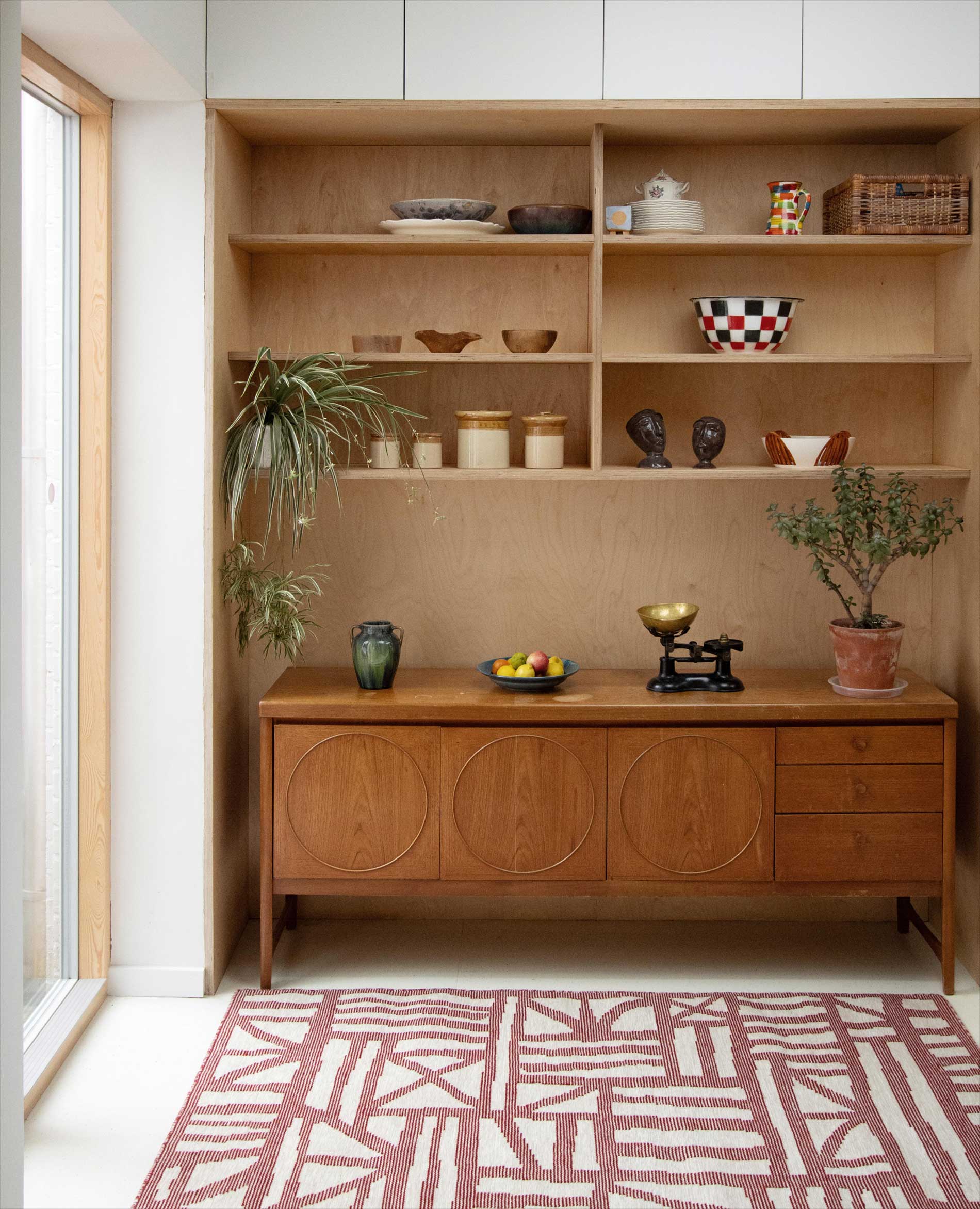 A wooden shelving unit with various decorative bowls, jars, plants, and a vintage scale stands against a wall. Below, a mid-century sideboard sits on a white floor with a red and white patterned rug. Light comes from a window on the left.