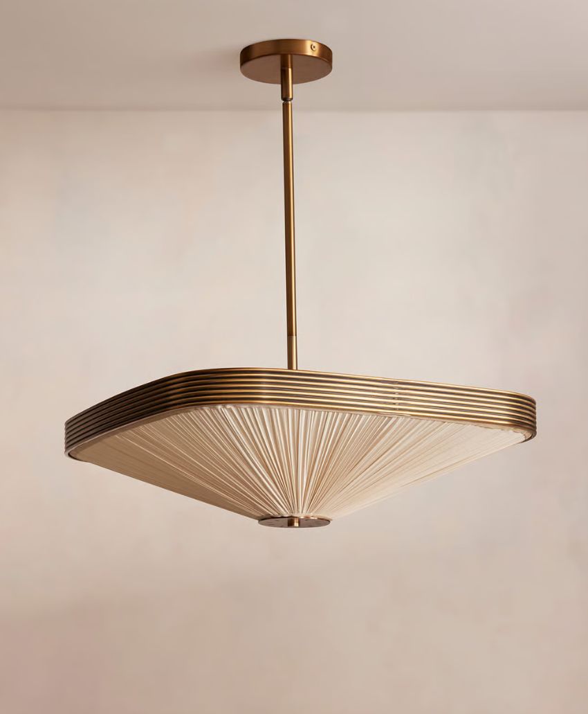 A modern pendant light with a brass fixture and a wide, pleated fabric shade, hanging from a ceiling against a neutral background.
