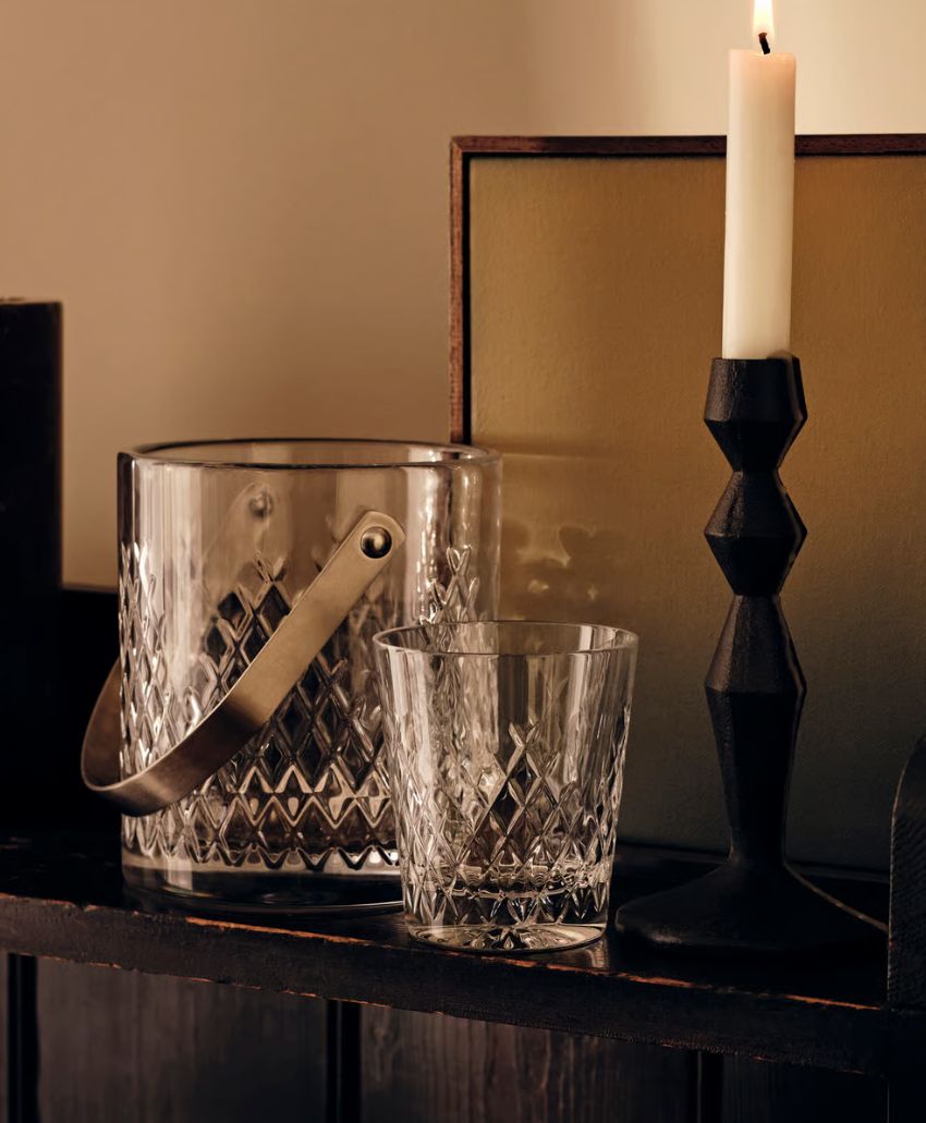 A lit white candle in a black geometric holder stands next to a cut-glass ice bucket and matching glass tumbler on a dark wooden shelf against a warm, softly lit background.