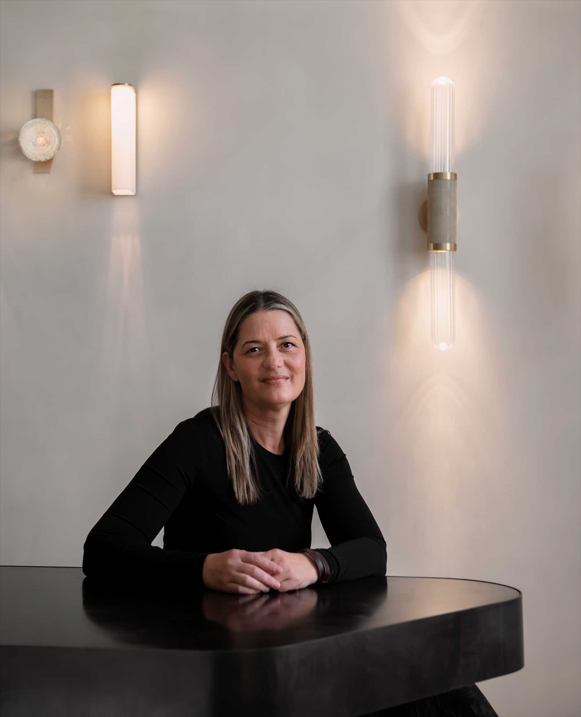 A woman with straight, shoulder-length hair sits at a dark table, smiling, with her hands folded. Three modern wall lights are illuminated on the pale wall behind her.