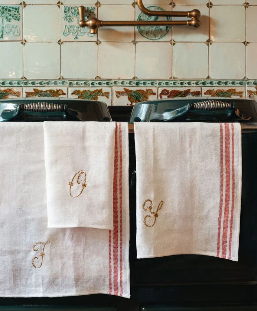 Two white tea towels with red stripes and embroidered initials hang on a black oven door. Behind them are cream-colored tiles with green patterns and a brass faucet above.