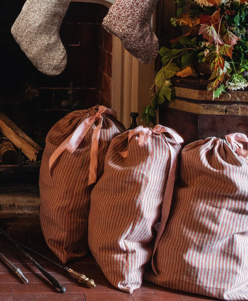 Three striped fabric sacks tied with ribbons are placed on a brick floor near a fireplace. A stocking hangs above, and greenery with autumn leaves decorates the scene, creating a cozy, festive atmosphere.