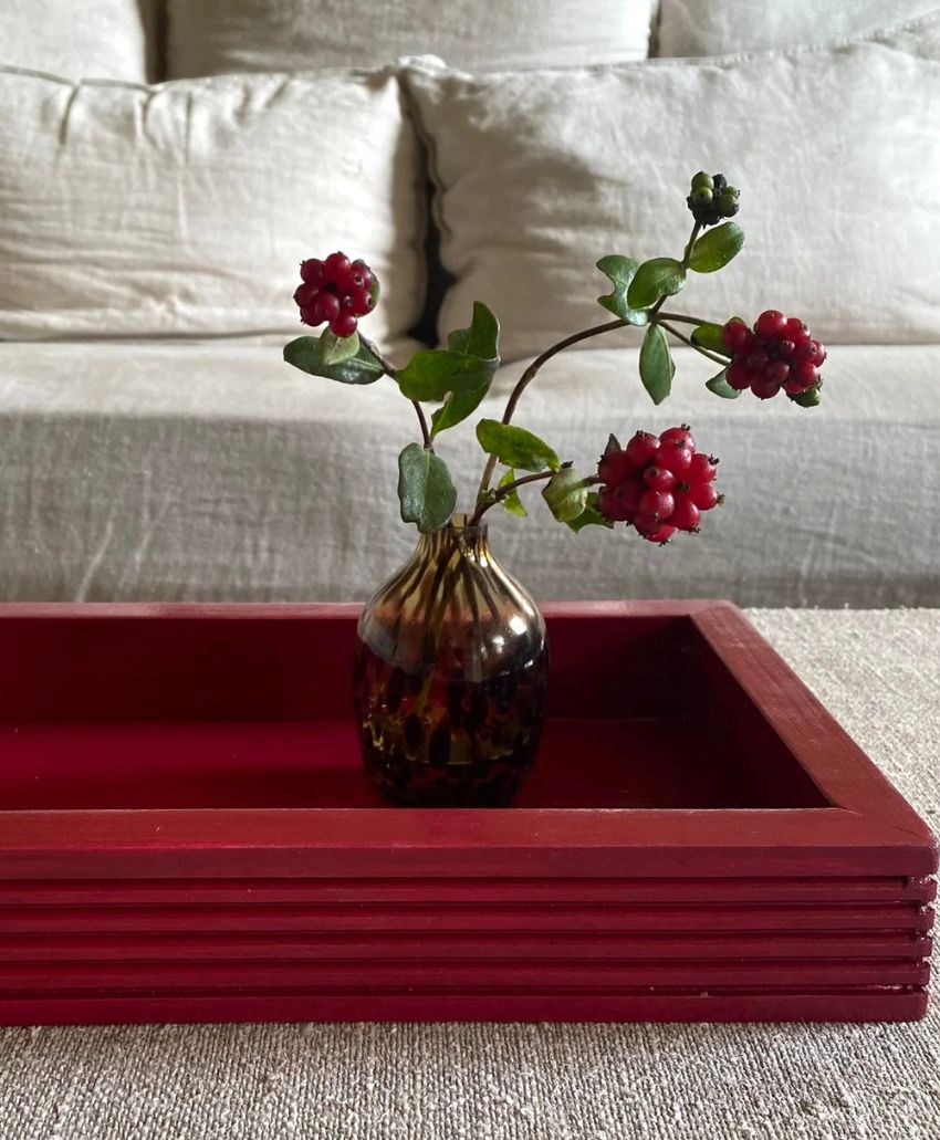A small glass vase with red flowers and green leaves sits in a rectangular red tray on a beige fabric surface, with a light-colored sofa in the background.