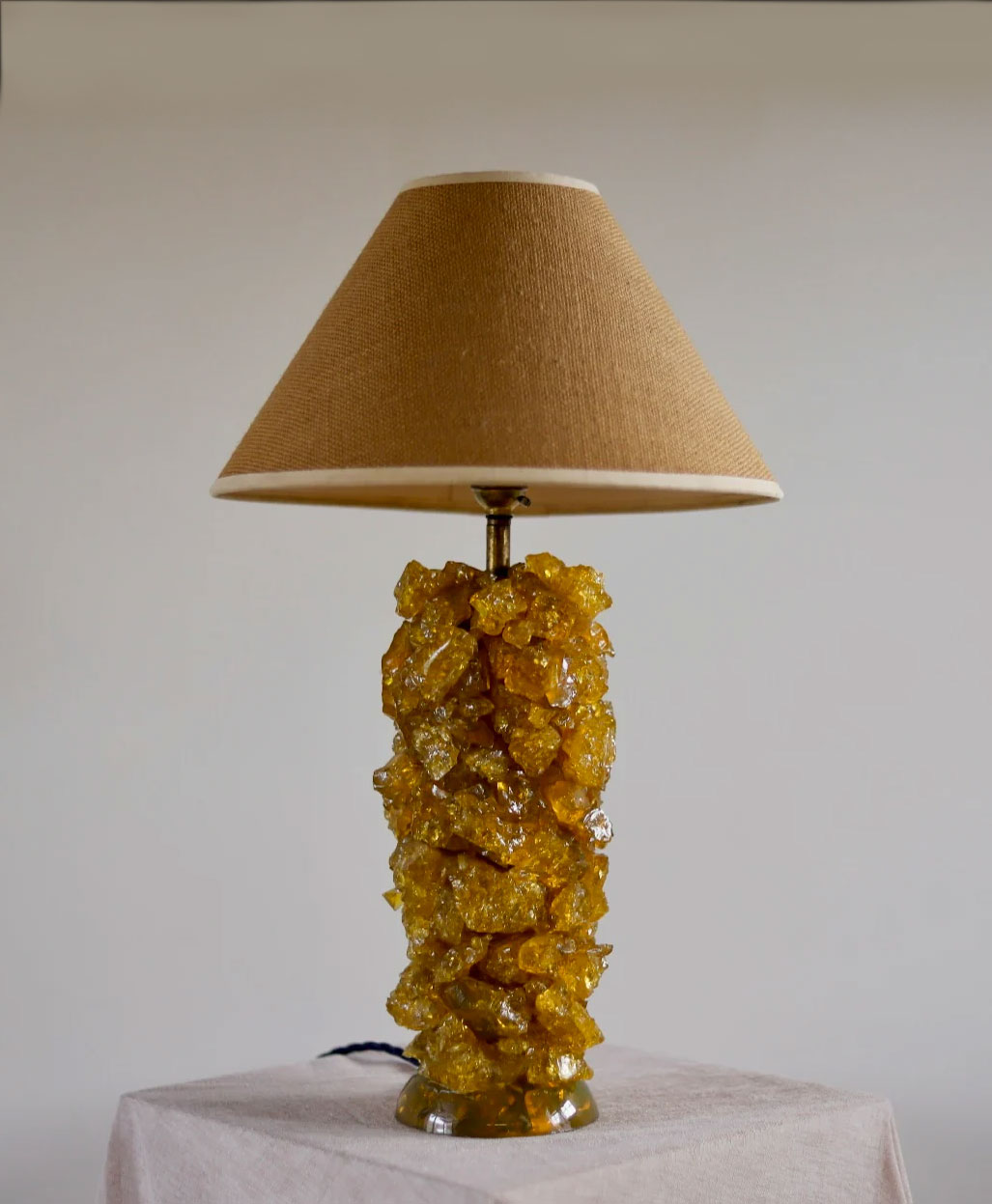 A table lamp with a tan fabric lampshade and a base made of clustered, textured yellow stones, sitting on a light-colored cloth-covered surface against a plain background.