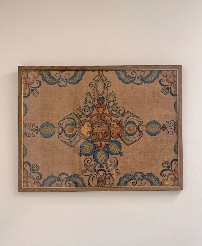 A framed, ornate artwork featuring a symmetrical, intricate pattern with blue, orange, and brown details on a beige background, hanging on a plain white wall.
