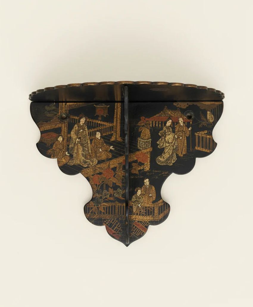A decorative black wooden wall shelf with scalloped edges, featuring intricate gold and red painted scenes of figures in traditional Asian attire within a garden or courtyard setting.