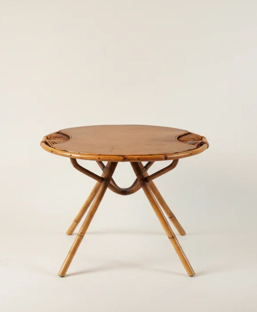 Round wooden side table with a smooth top and three angled, interlaced legs, set against a plain, light-colored background. The table has a natural, rustic appearance with visible wood grain.