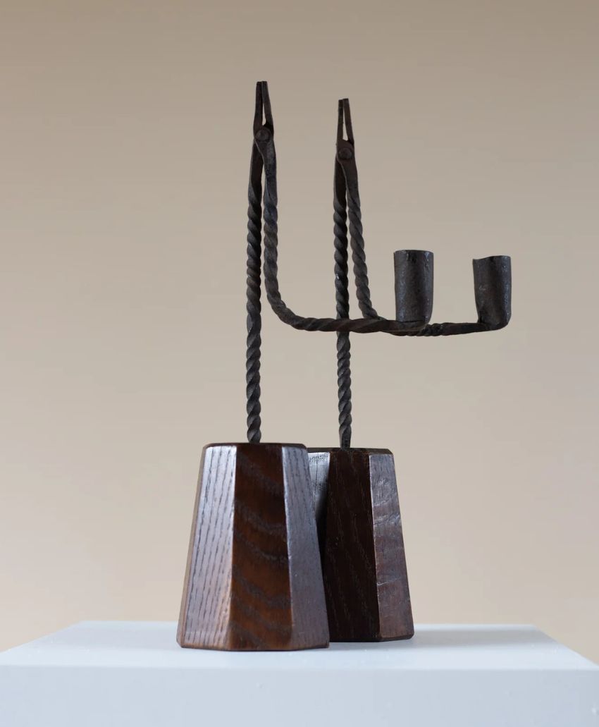 A sculptural object made of twisted metal and dark wood. Two vertical metal rods with small cup-like ends rise from angular wooden bases, forming an abstract, candelabra-like figure on a white pedestal.