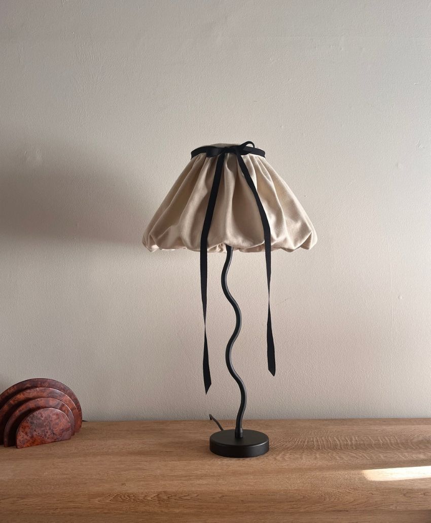 A lamp with a wavy black stand and a cream-colored, fabric lampshade tied with black ribbons sits on a wooden surface against a plain light-colored wall. Marble coasters are stacked on the left.