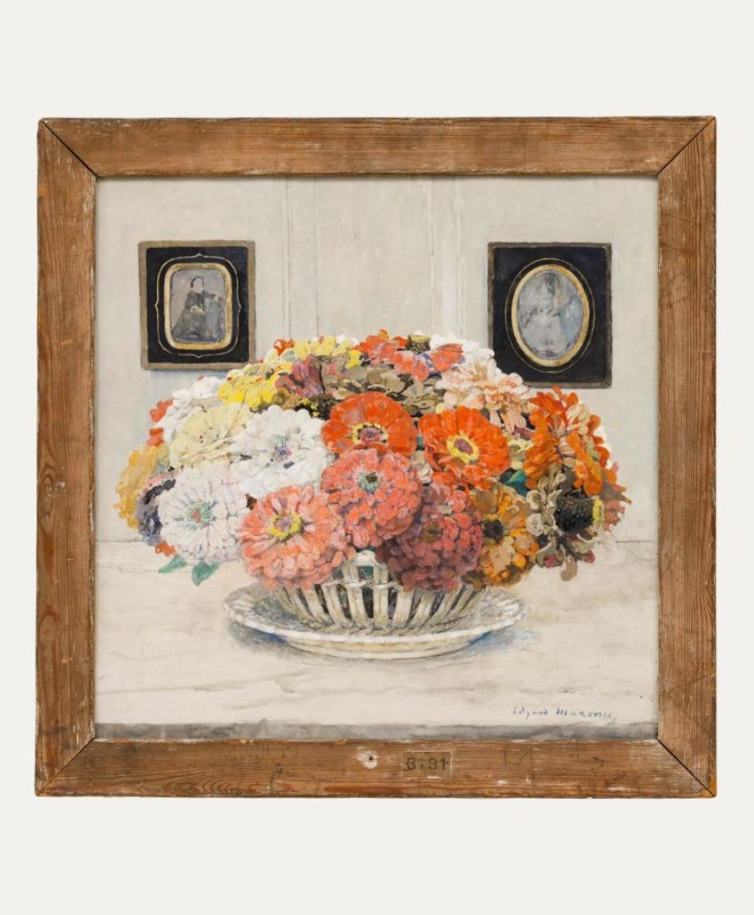 A framed painting shows a basket filled with colorful flowers on a white table. In the background, two small, vintage portraits hang on a plain, light-colored wall.
