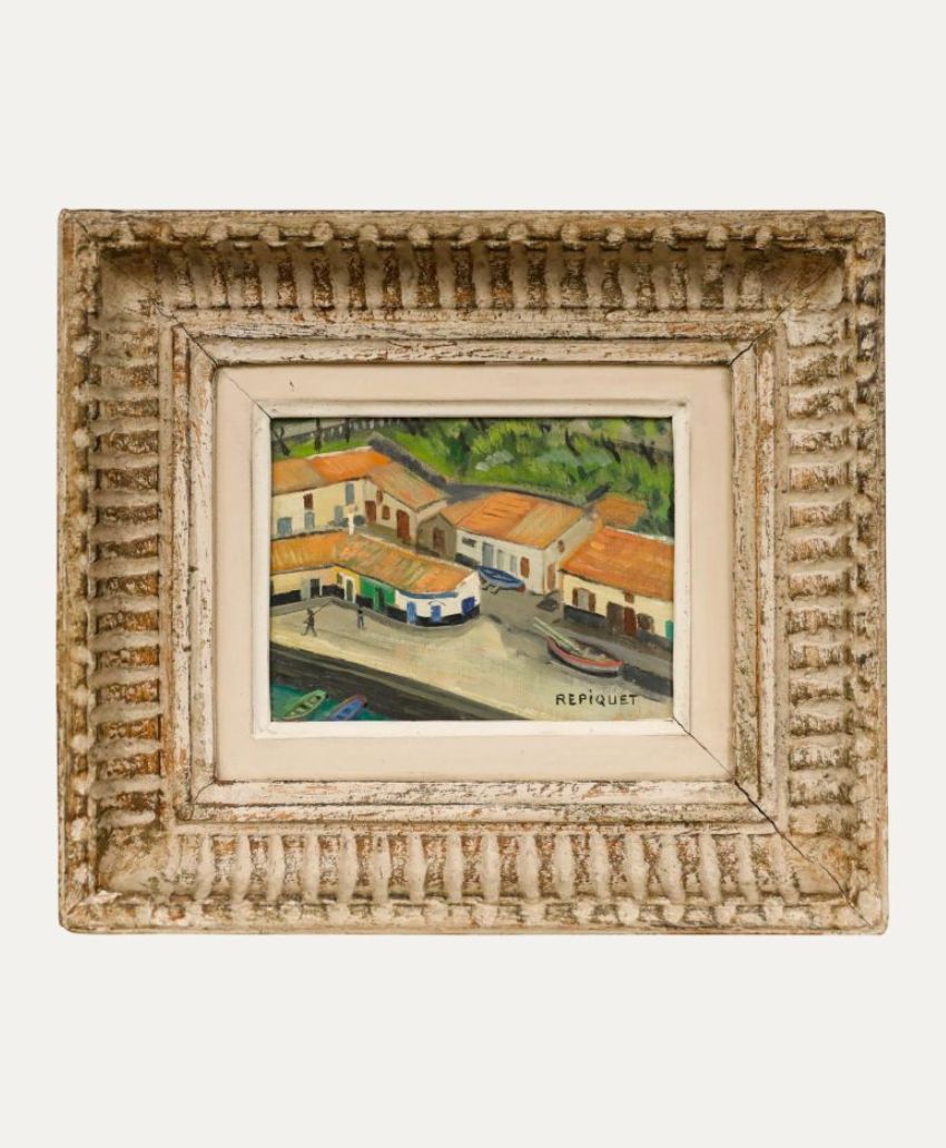 Framed painting of a street scene with white buildings, red roofs, green trees, and people walking; signed R. Piquet. The frame is ornate and rustic with a textured, weathered finish.