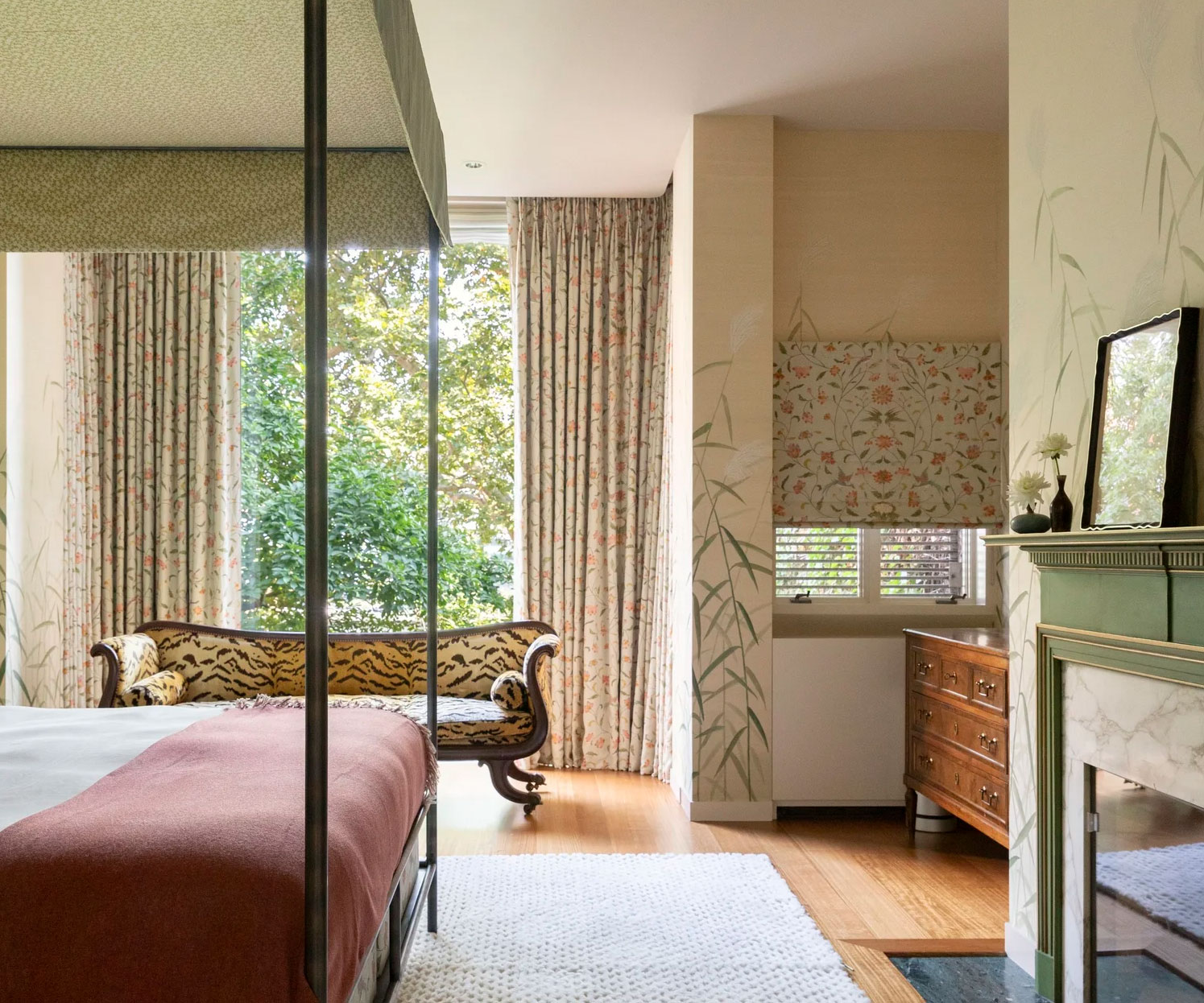 A bright, elegant bedroom with large windows, floral curtains, a four-poster bed, a vintage wooden sofa with animal print, a wooden dresser, and a decorative fireplace with a mirror above it.