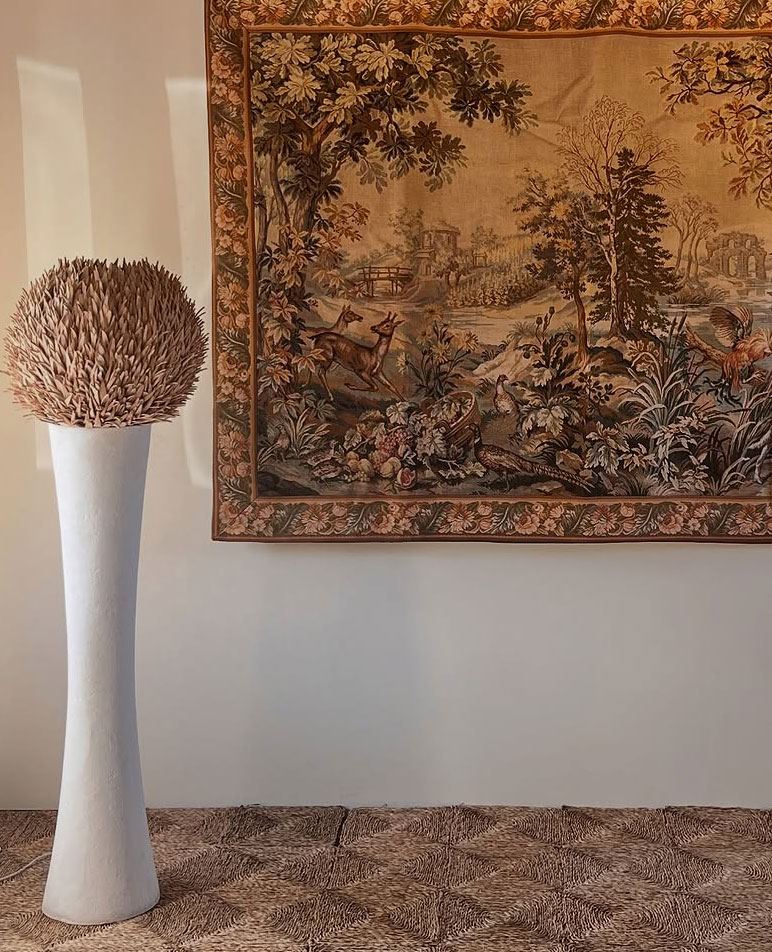A tall, white textured vase with a feathery arrangement stands next to a wall adorned with a detailed tapestry depicting a forest scene with deer, trees, and foliage in warm, earthy tones.