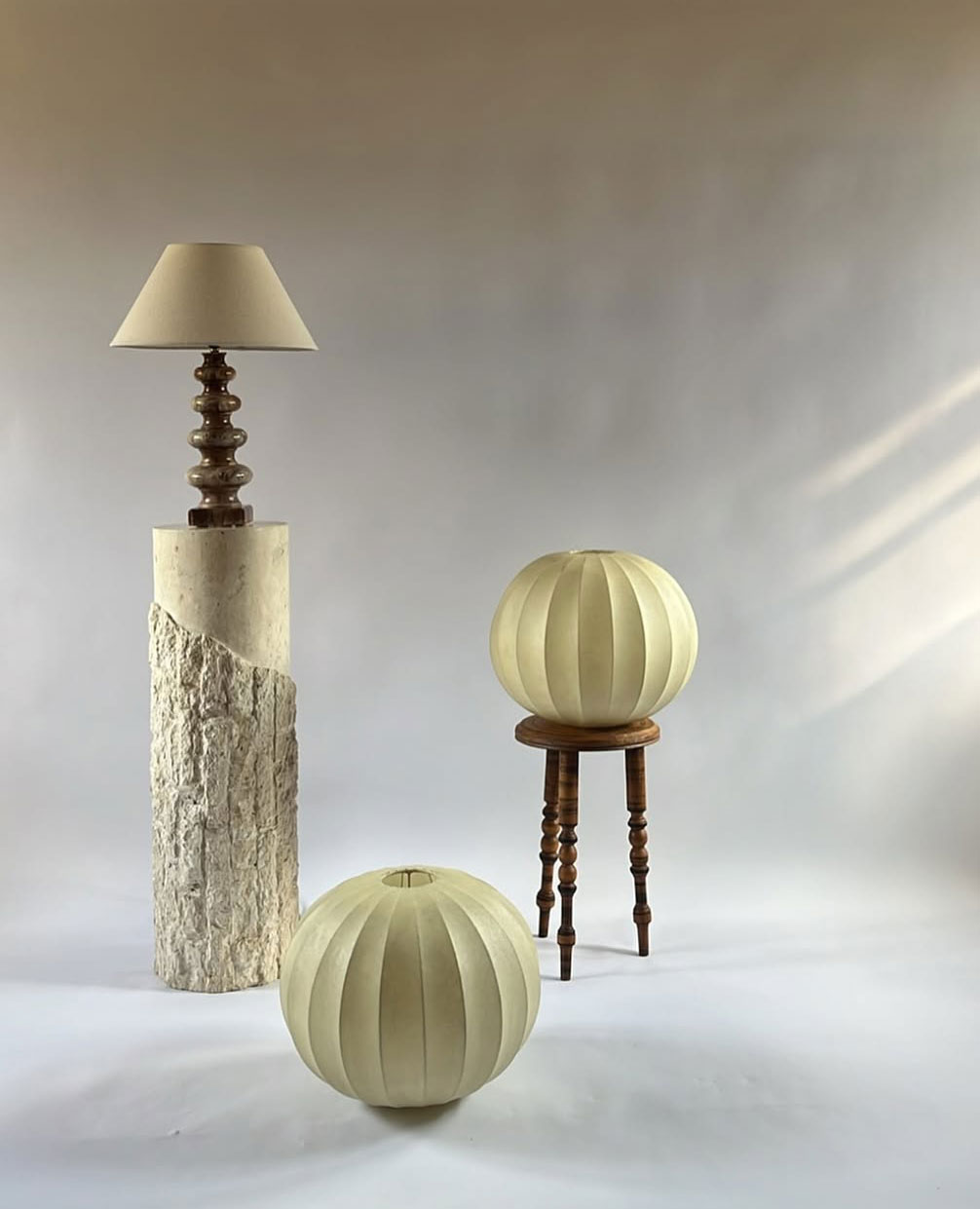 Three unique lamps: one with a beige shade on a textured stone and wood base, and two round lamps with pleated off-white shades—one on the floor, one on a wooden stool—set against a neutral background.