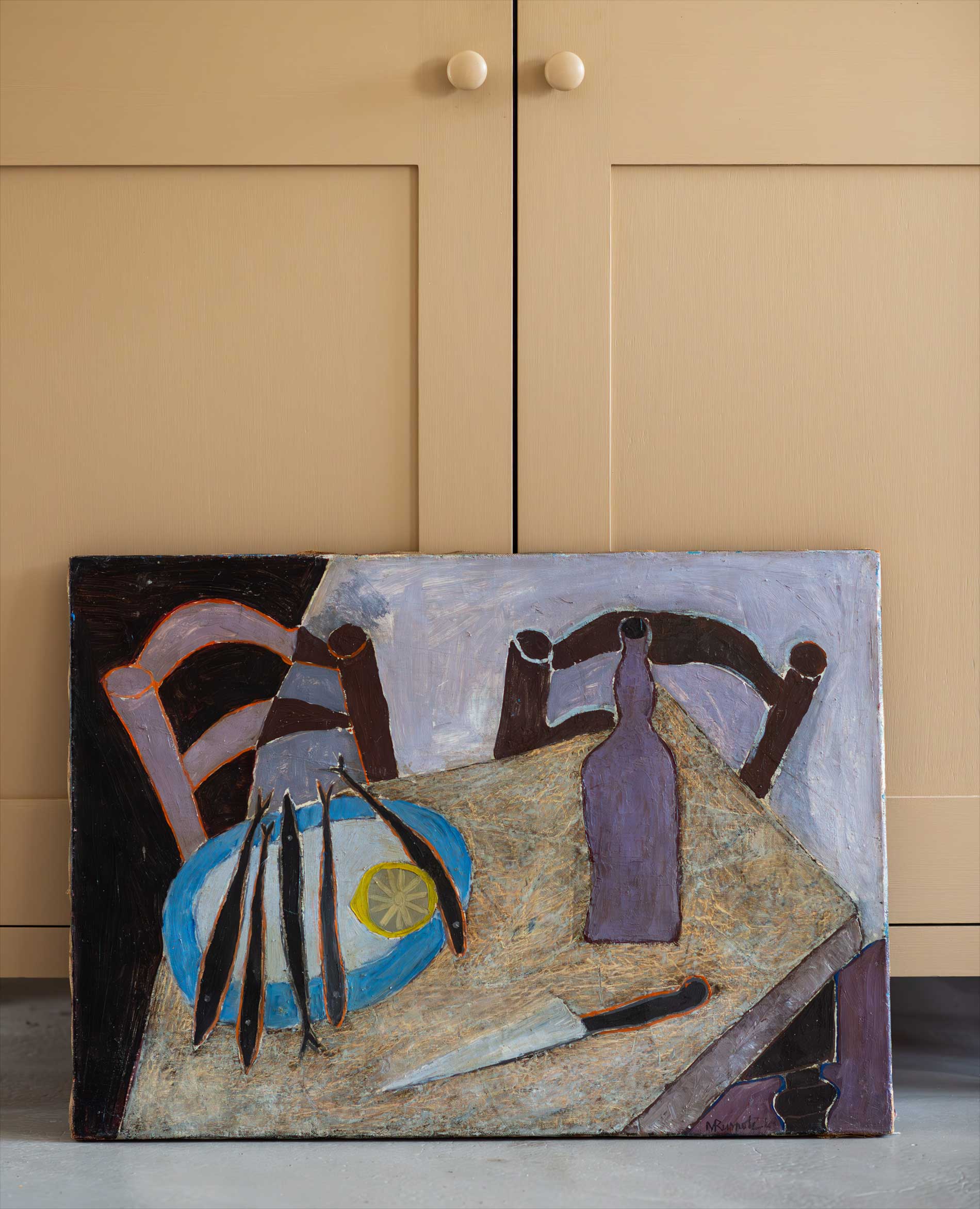 A painting leans against beige kitchen cabinets. The painting depicts a table with a blue plate holding fish and a lemon slice, a knife, and a purple bottle. Two wooden chairs are partially visible in the background.