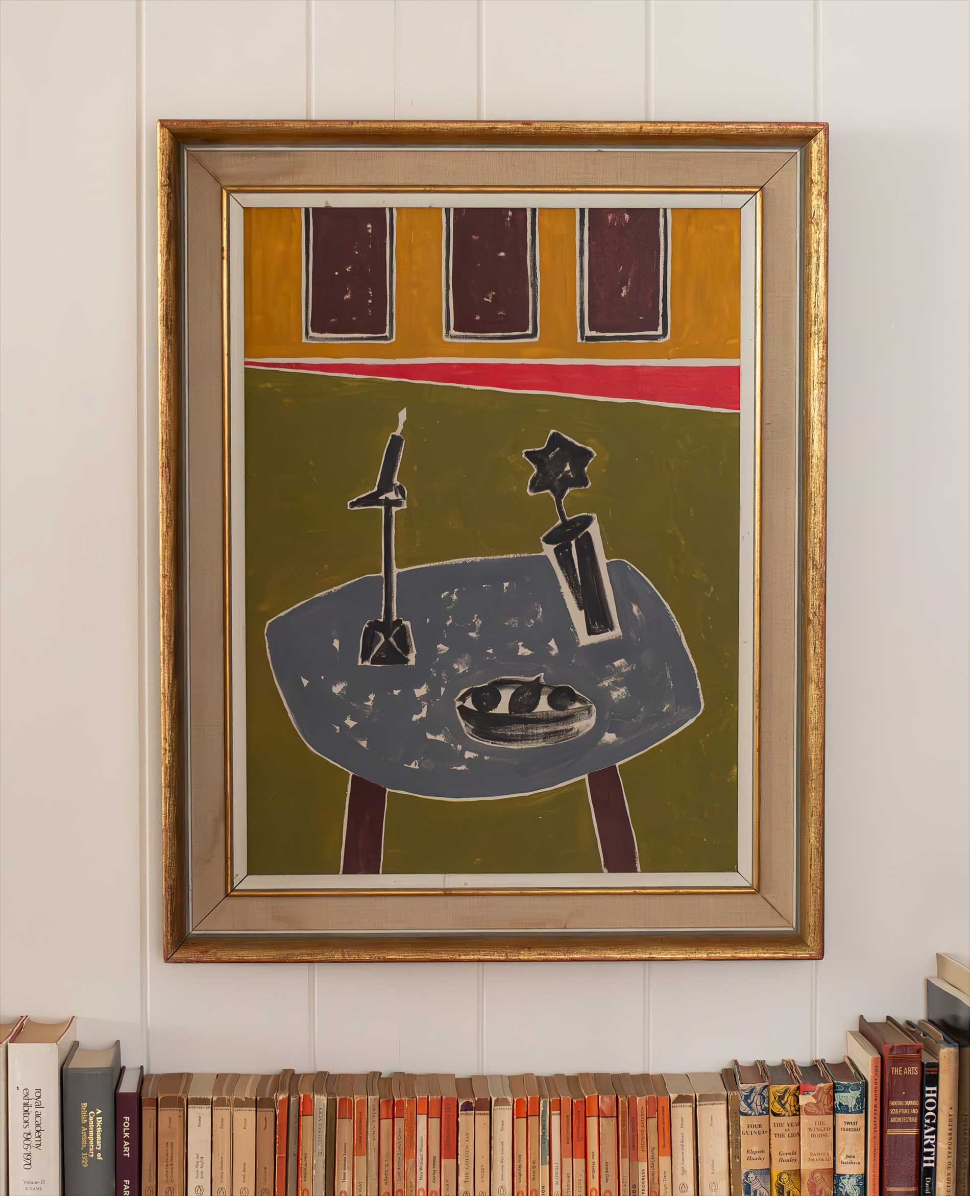 A framed abstract painting hangs on a white wall above a shelf of books. The artwork features a table with geometric objects and a dish, set against a background with rectangles and diagonal lines in earthy tones.