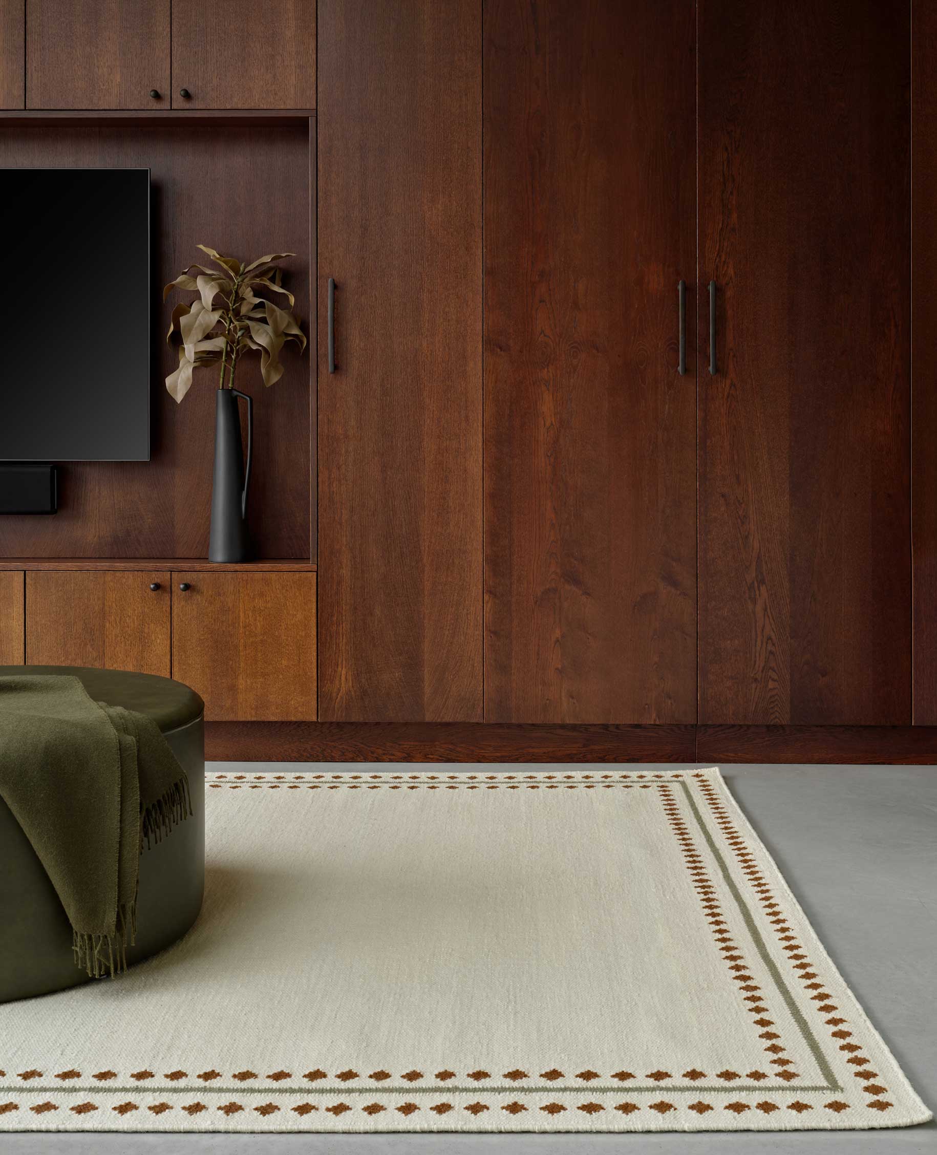 A modern living room with dark wood cabinetry, a mounted flat-screen TV, a decorative vase with dried foliage, an olive green ottoman, and a light rug with a geometric border pattern.