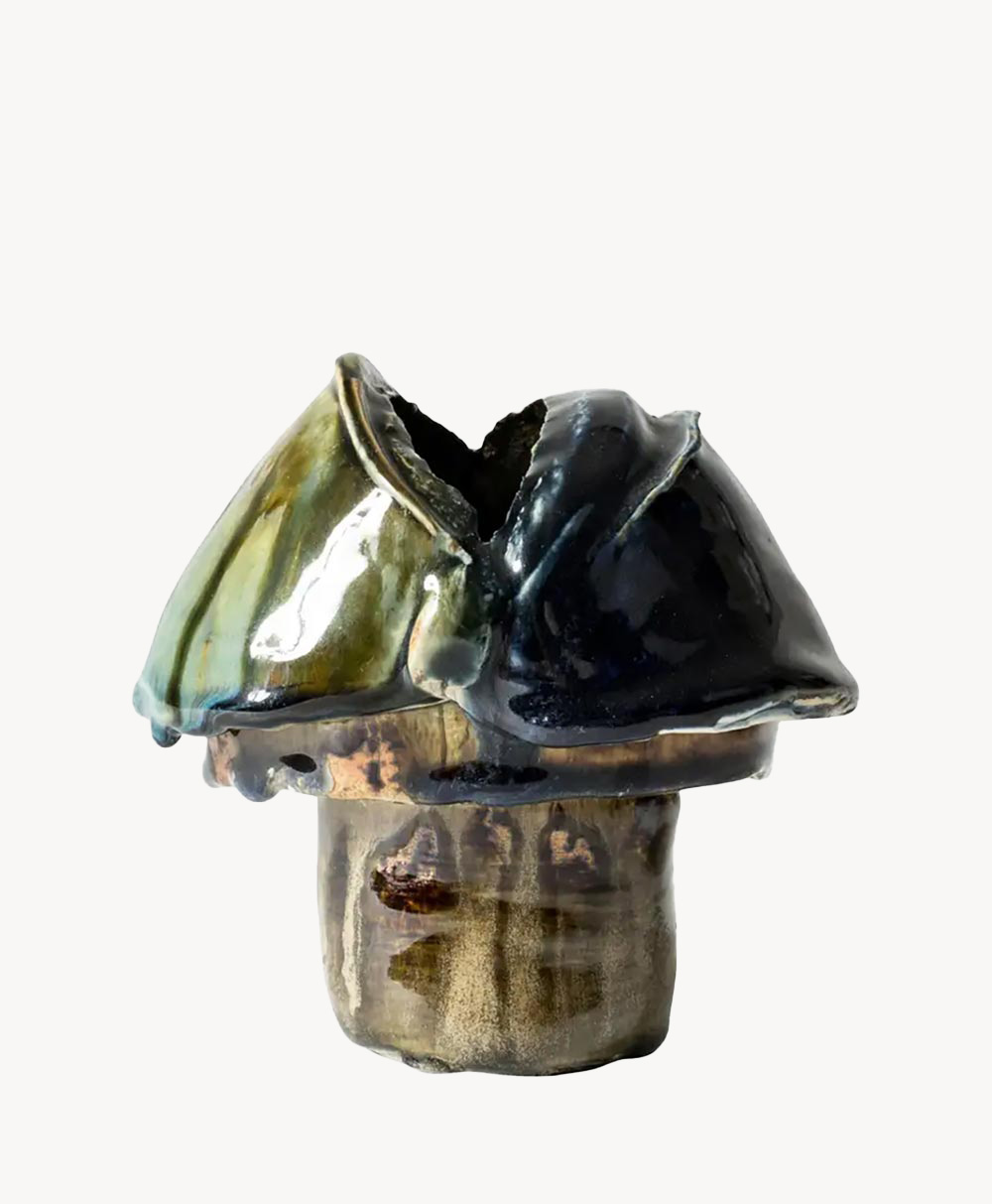 A ceramic sculpture resembling a mushroom with a thick stem and a glossy, irregularly shaped cap in shades of dark blue, green, and brown. The artwork has a textured and organic appearance.