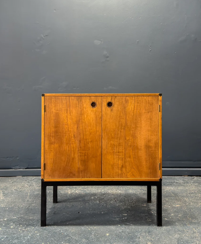 A small, mid-century modern wooden cabinet with two doors and circular cut-out handles, standing on black legs against a dark gray wall and concrete floor.