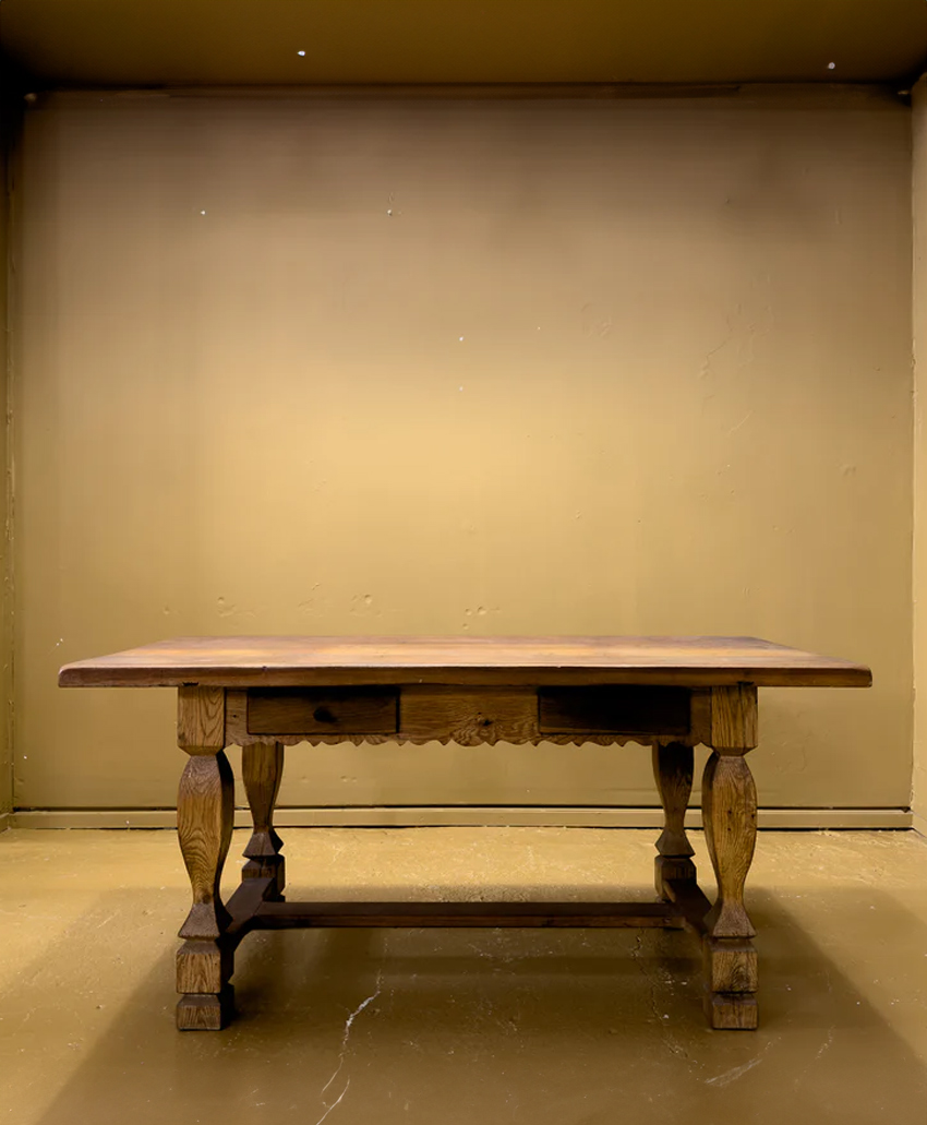 A wooden table with ornate legs and a carved apron stands against a plain, beige-colored wall and floor in an empty, well-lit room.