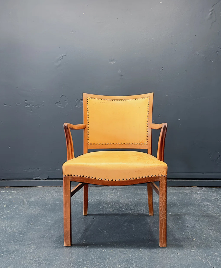 A wooden chair with a yellow upholstered seat and backrest, featuring armrests and decorative brass studs, sits against a plain dark gray wall on a dark floor.