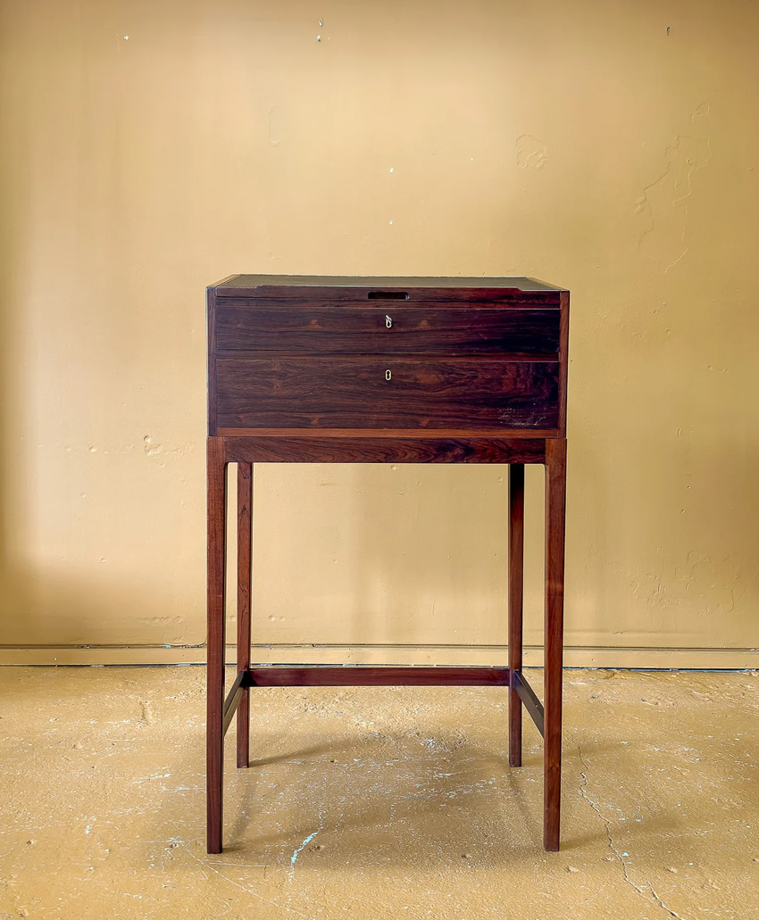 A dark wooden box with two drawers stands on tall, slender wooden legs against a plain beige wall and floor. The box has simple, clean lines and a minimalist design.