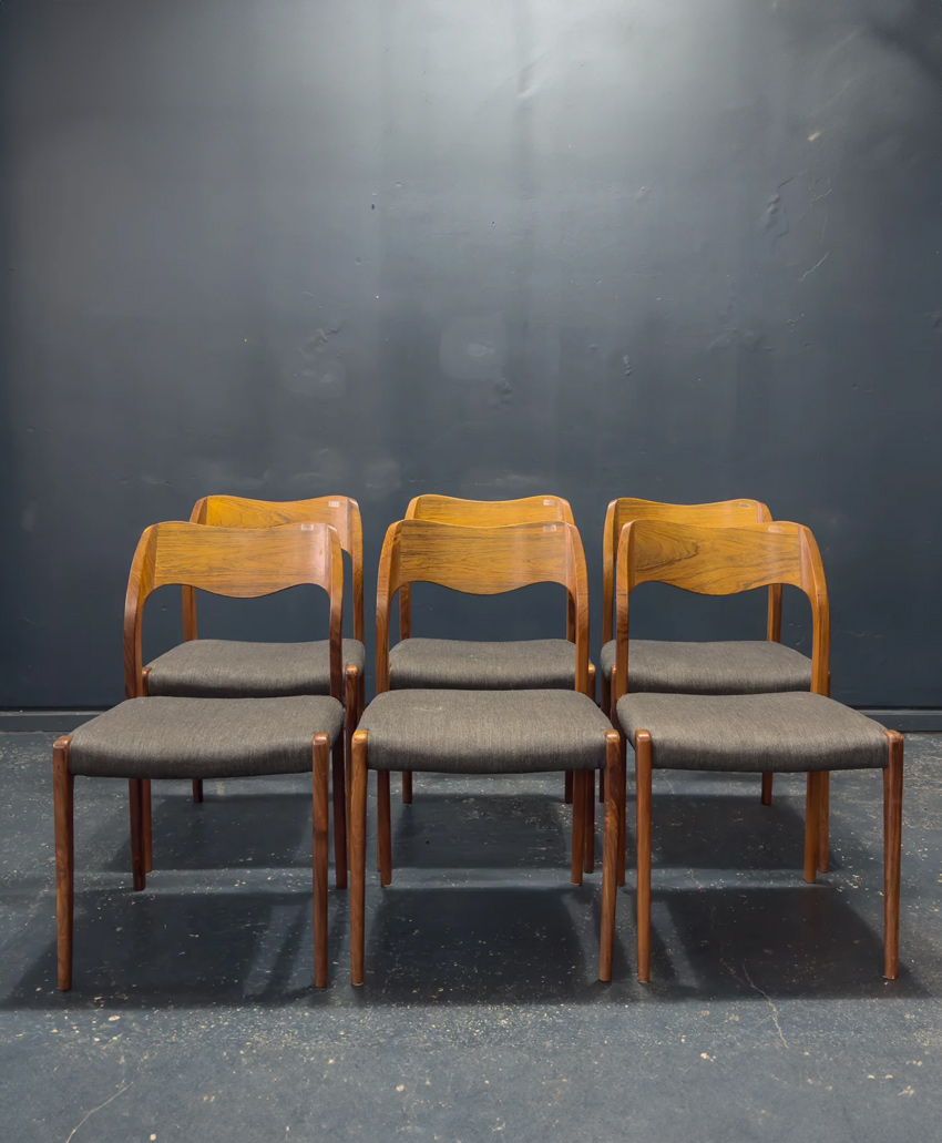 Six mid-century modern wooden chairs with curved backs and dark upholstered seats are arranged in two neat rows of three, set against a plain, dark background and floor.