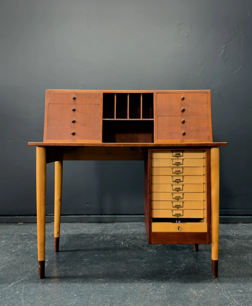A vintage wooden desk with slender legs, small drawers on the left and upper sections, open cubby slots in the center, and a vertical row of tan drawers on the right side, set against a dark gray wall.