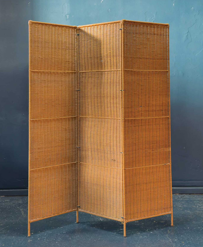 A four-panel folding room divider made of woven rattan stands on a dark floor against a blue wall. The panels are connected by hinges, allowing the screen to bend into a zigzag shape.