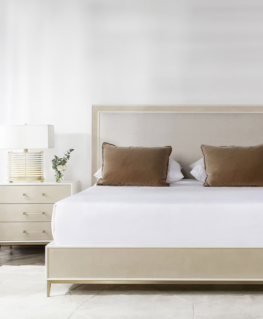 A modern, minimalist bedroom with a beige bed frame, white bedding, two large brown pillows, a white nightstand with a lamp, and a small plant on the side. The walls are plain white.