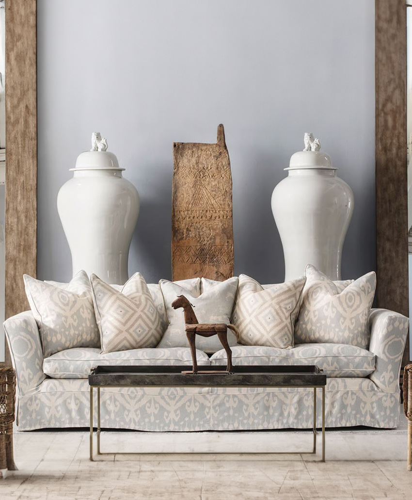 A living room features a patterned sofa with neutral pillows, a glass coffee table holding a wooden horse sculpture, two large white ceramic vases, and a carved wooden panel against a light gray wall.