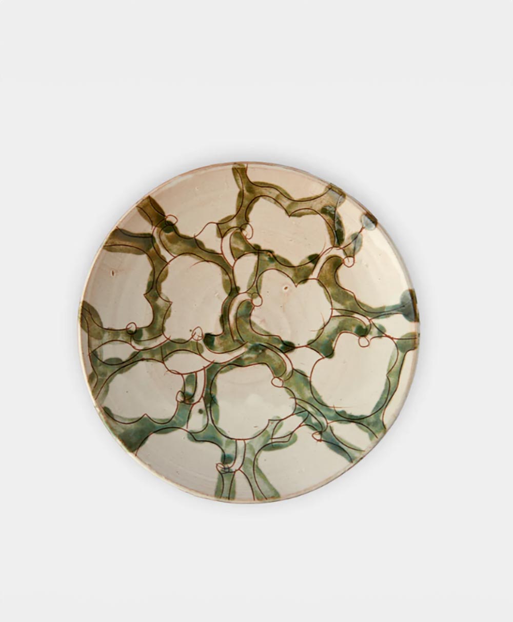 A round ceramic plate with an abstract, organic pattern of green and brown lines and shapes on a beige background, resembling interconnected natural forms. The plate is displayed against a plain white surface.