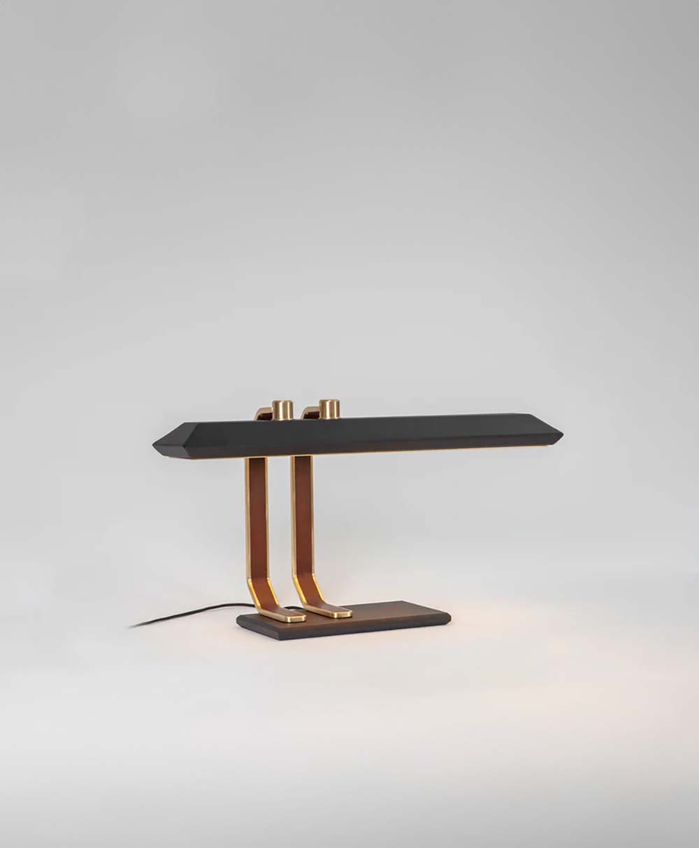 A modern desk lamp with a sleek black rectangular shade, gold-colored double support arms, and a matching rectangular base, set against a plain light gray background.