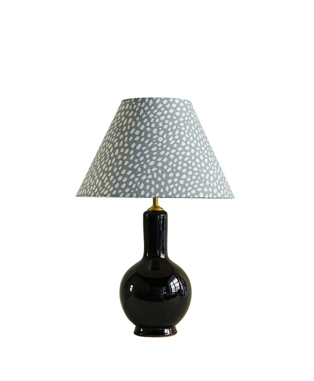 A table lamp with a glossy black, round base and a wide, tapered lampshade featuring a light gray background with white speckled dots, set against a plain white background.