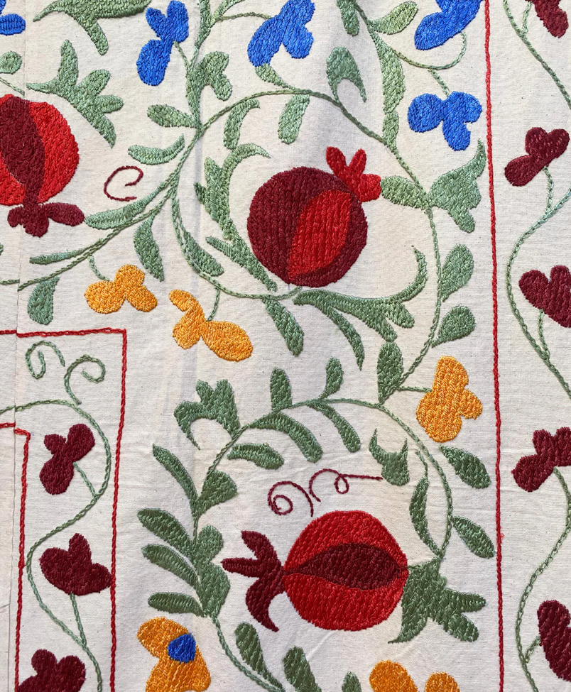 Colorful embroidery featuring red pomegranates, blue and yellow flowers, and green leaves on a white fabric background, with swirling vine patterns and decorative detailing.