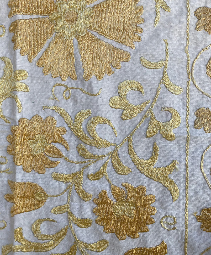 Gold floral embroidery on white fabric, featuring intricate flowers and vines with dense, textured stitching, creating an ornate and decorative pattern.