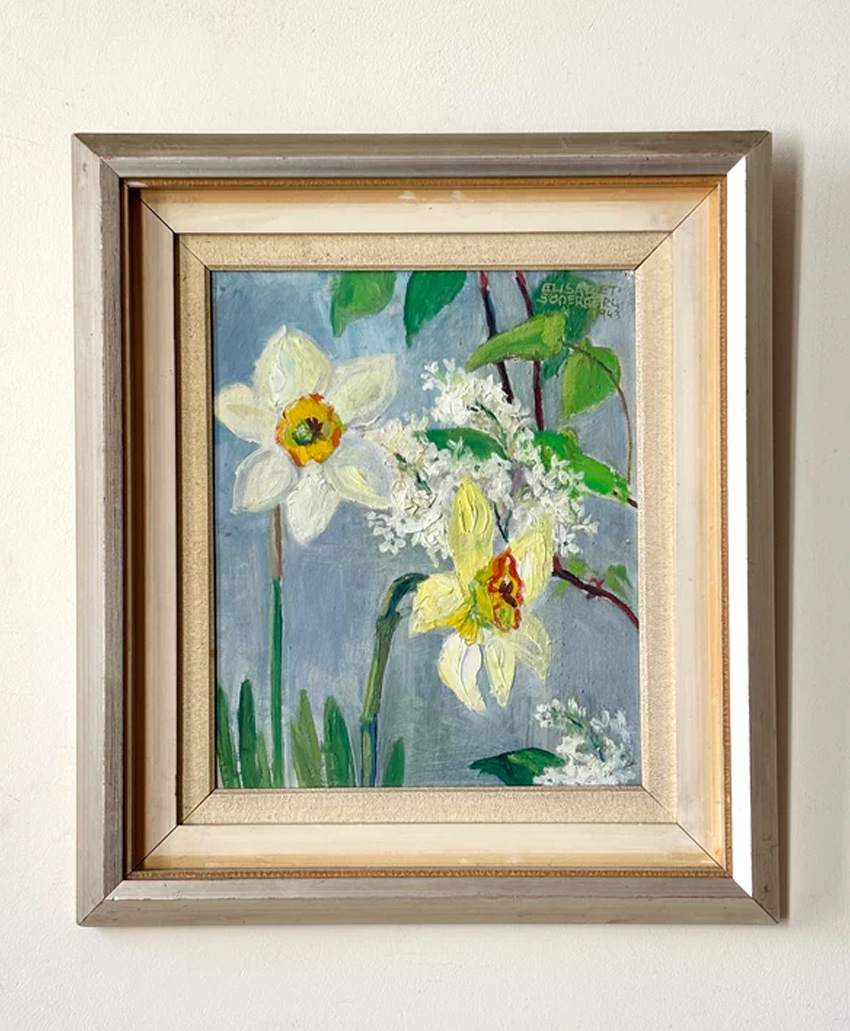Framed painting of white and yellow daffodils with green stems and leaves against a blue-gray background, hung on a plain white wall.