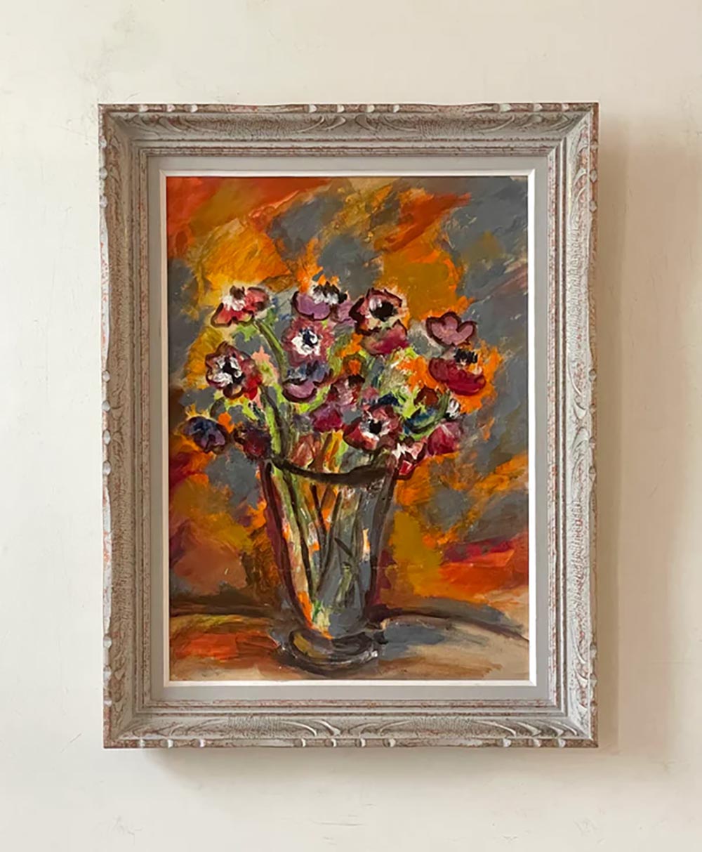 A framed painting of a vase filled with vibrant, colorful flowers against an abstract orange, yellow, and gray background, hanging on a light-colored wall.