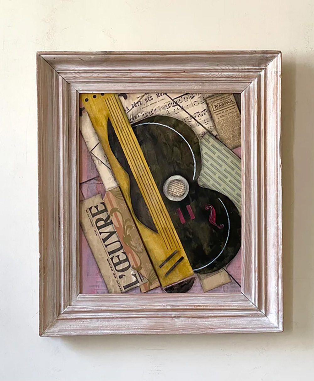 Framed abstract artwork featuring a stylized guitar, newspaper clippings, and sheet music with muted colors and textured layers, displayed against a light-colored wall.