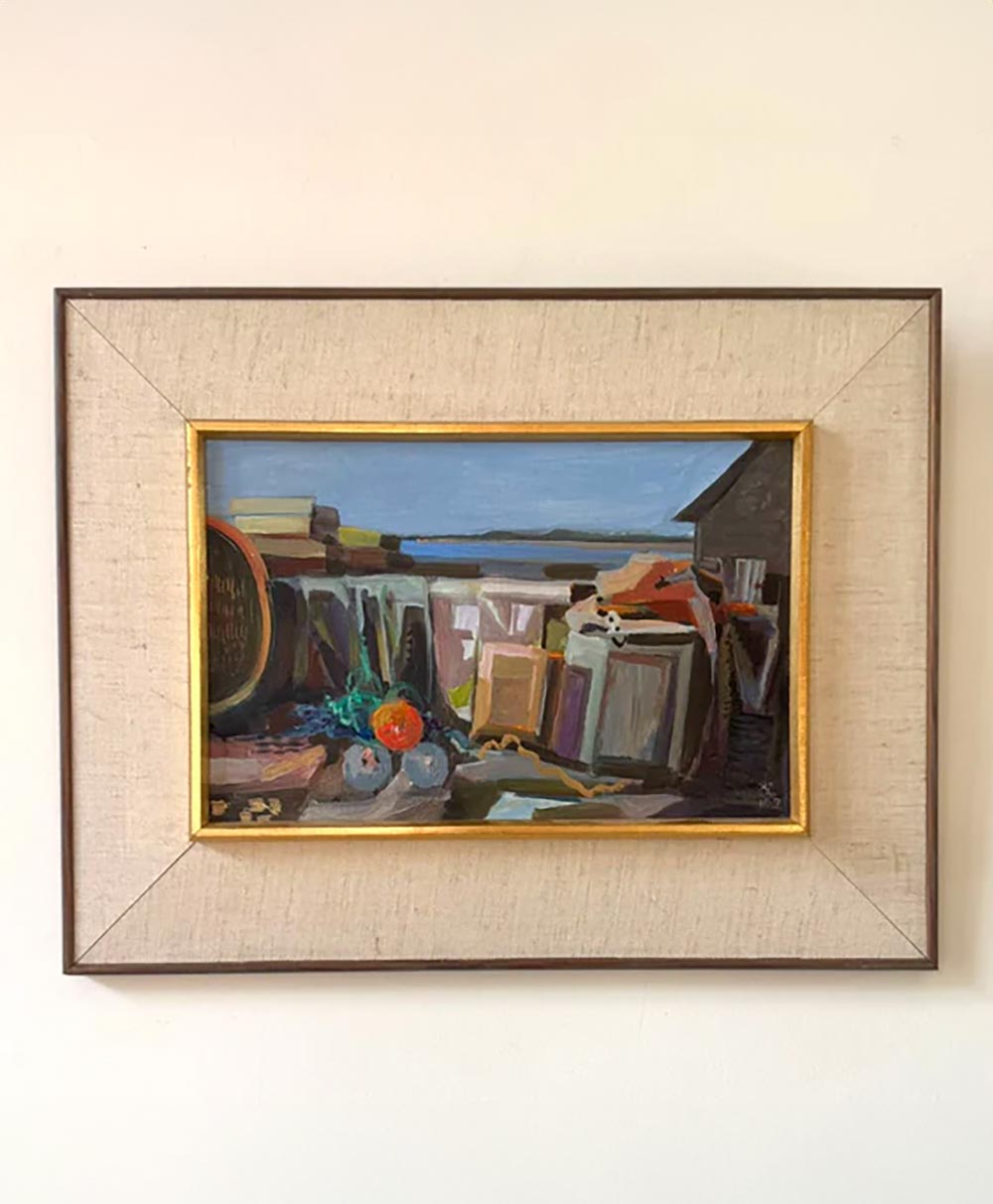 A framed painting showing a still life with books, a bottle, some fruit, and small objects on a table, set against a blue sky and building background, hanging on a plain white wall.