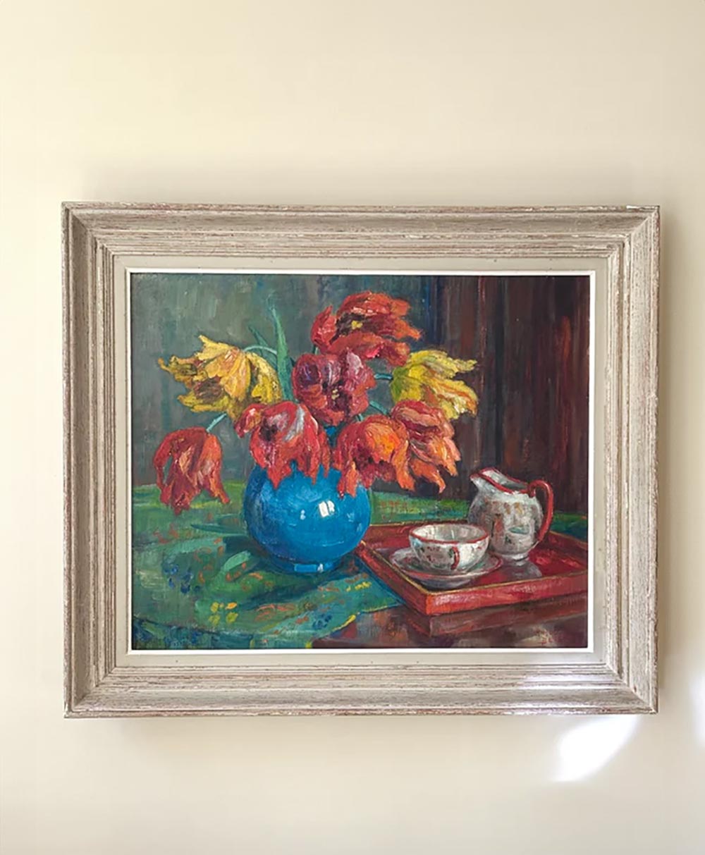 Framed painting of wilted red and yellow flowers in a blue vase, with a teacup and creamer on a red tray beside them, set against a green tablecloth and a muted background.
