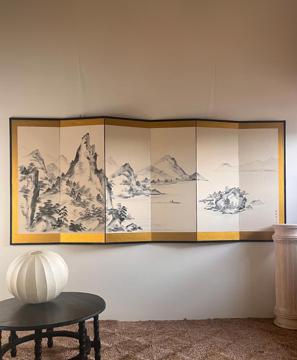 A Japanese folding screen with a black-and-white mountain landscape painting hangs on a wall above a round black table with a white vase and a beige column pedestal, on a brown carpeted floor.