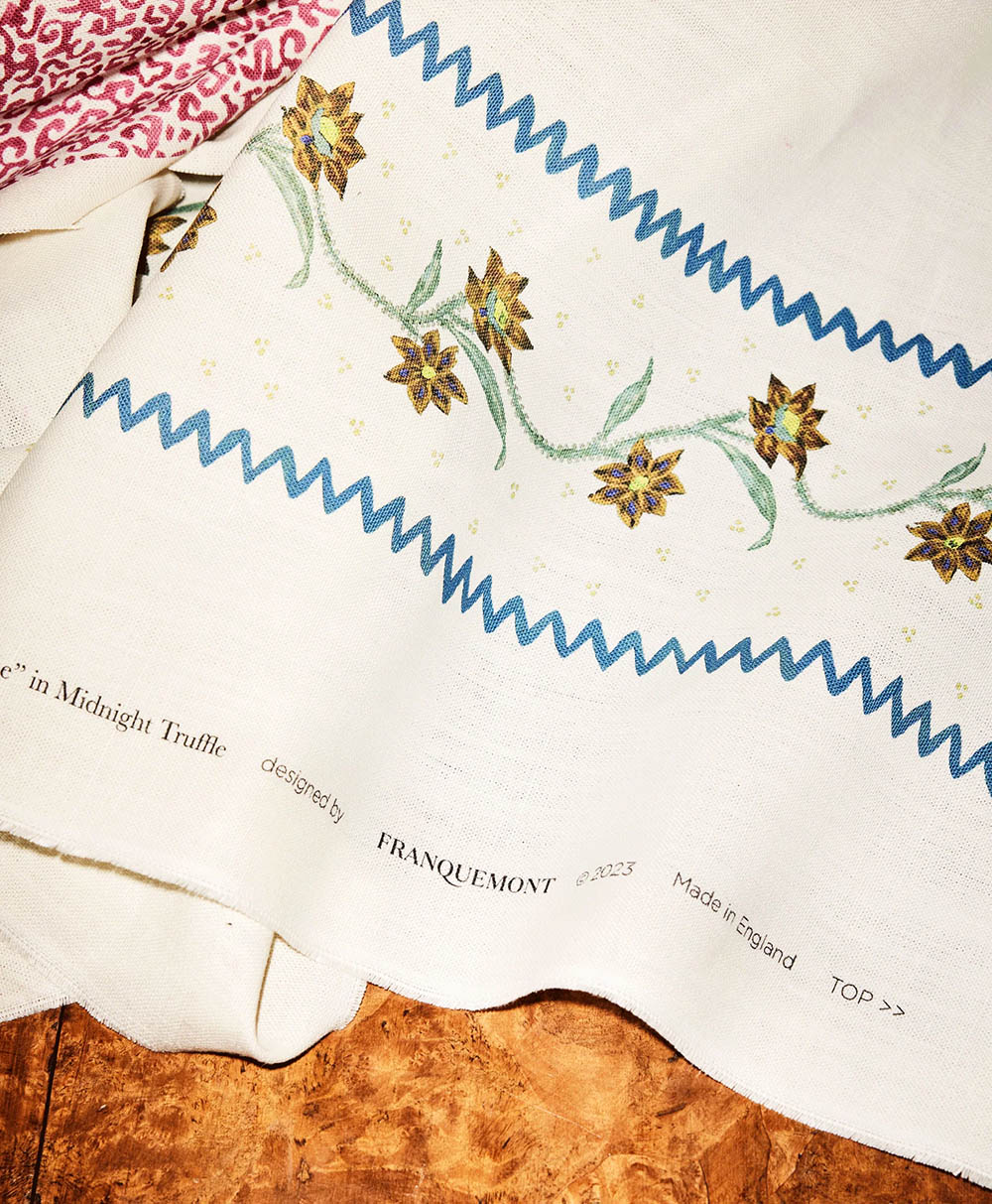 A close-up of a cream-colored fabric with a blue zigzag border and a floral vine pattern. Text along the edge reads “designed by FRANQUEMONT,” “Made in England,” and other small print. A red-patterned fabric is partially visible.